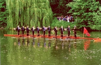 A photo of the 'Monarch' next to a riverbank with a crowd looking on. All of the rowers are standing up.