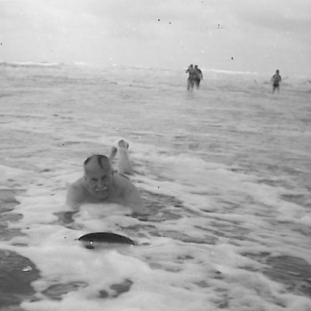 A black and white photo of Geoffrey Knight using a 1920s wooden bodyboard in shallow waters.