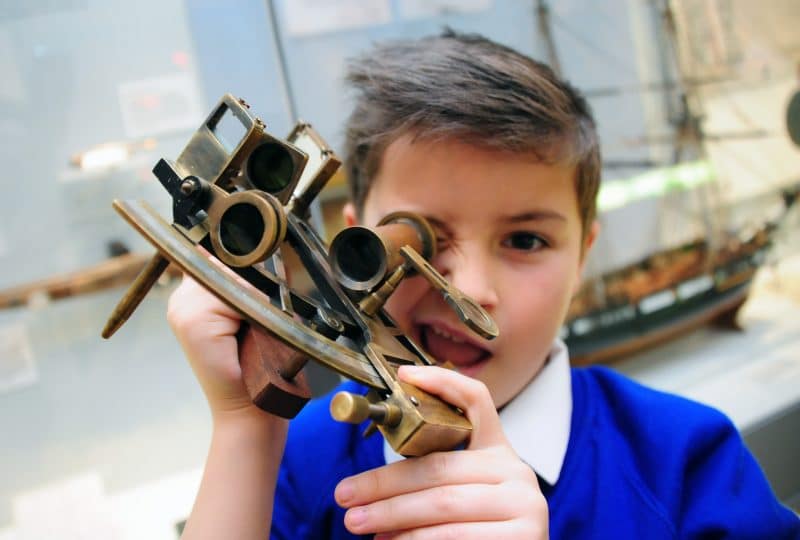 A photo of a schoolboy looking at the camera through a sextant that he holds up to his eye.