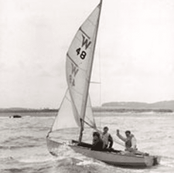 A black and white photo of three people sailing a Wayfarer dinghy. One man waves towards the camera.