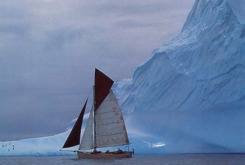 Photo of the 'Curlew' sailing in front of a very large glacier, with some penguins on the snow in the background to the left.