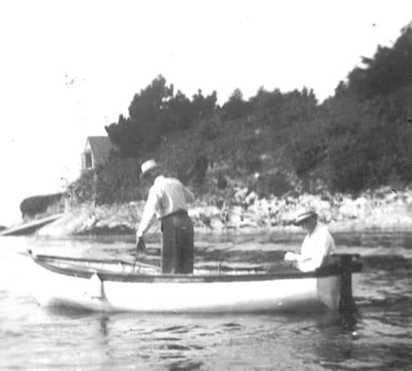 A black and white photo of two men sailing in the 'White Owl'. One is seated at the stern, the other is standing towards the bow.