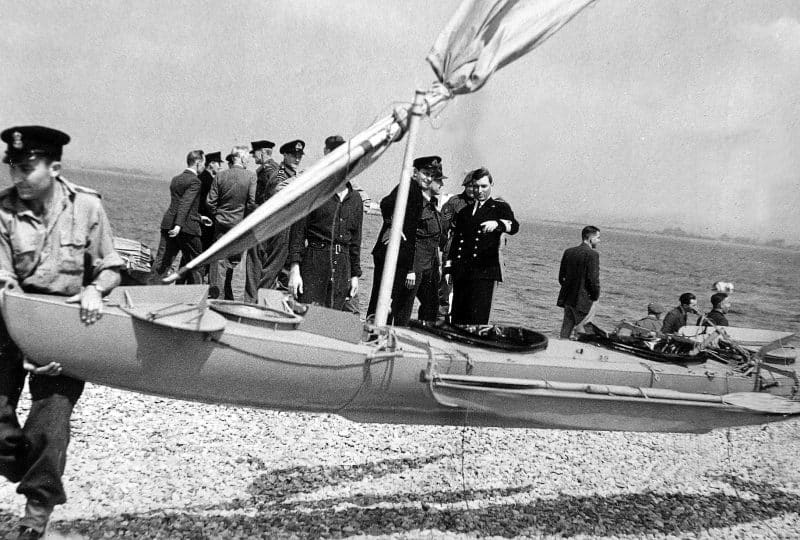 Black and white photo of the Mk.VII Canoe being carried up a beach. Behind it is a group of men in naval uniforms.