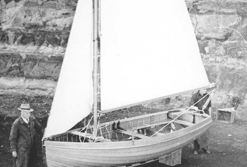 A black and white photo of the RNSA dinghy. One man stands at the bow, and another stands at the stern.