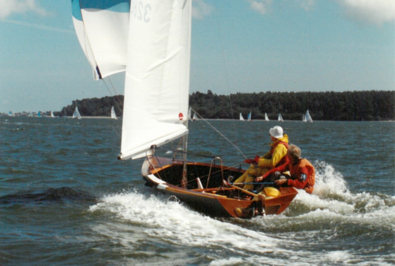 A photo of two men sailing 'The Feet', one wearing an orange sailing suit and the other wearing a yellow one with a white hat.
