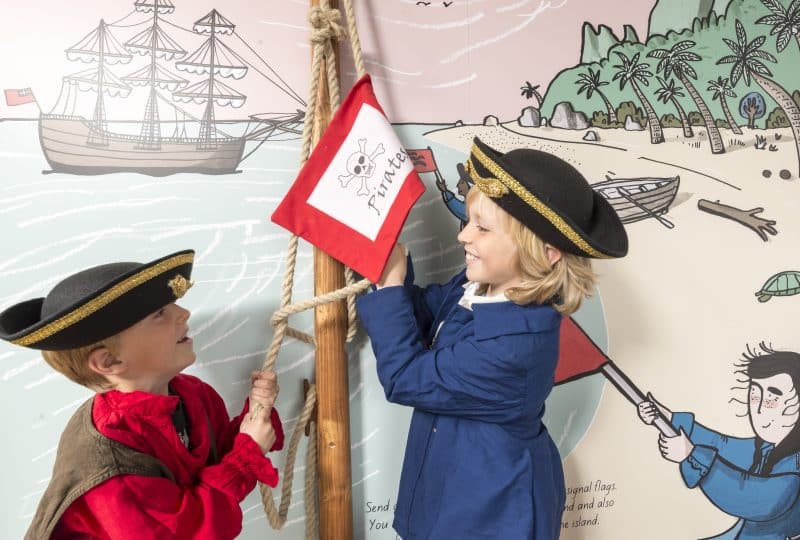 A photo of two young boys wearing sailors' hats, hoisting a flag with a skull and crossbones on it up a small flagpole.