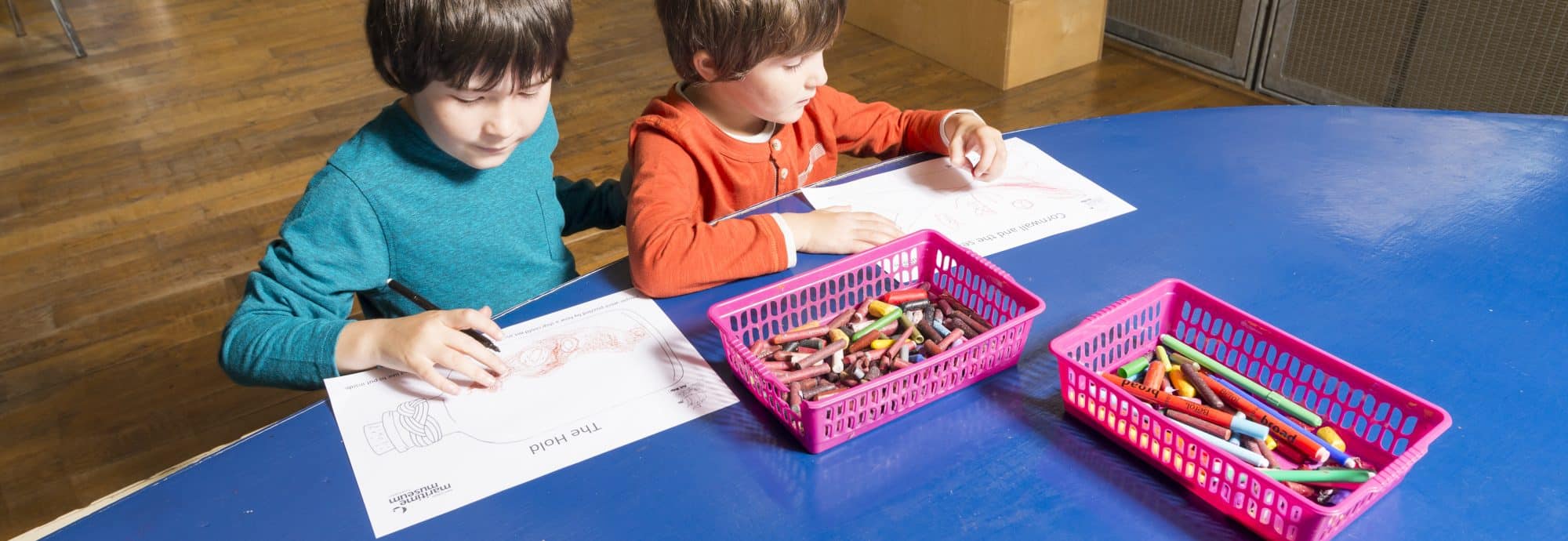 A photo of two young boys drawing as part of craft activities at the Museum. Two boxes of pens and crayons are in front of them on the blue table.