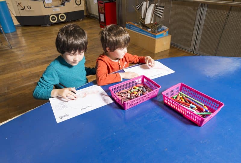 A photo of two young boys drawing as part of craft activities at the Museum. Two boxes of pens and crayons are in front of them on the blue table.