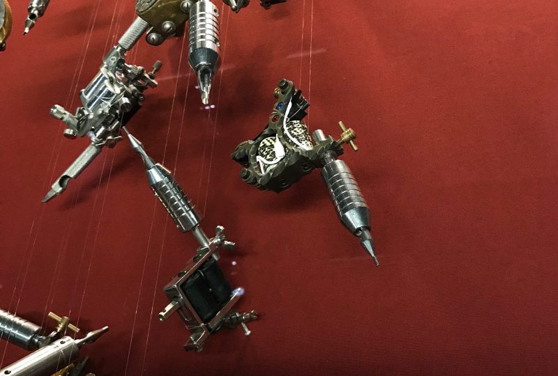 A photo of a cascade of parts of tattoo guns suspended against a red background as part of the Museum's 'Tattoo: British Tattoo Art Revealed' exhibition.