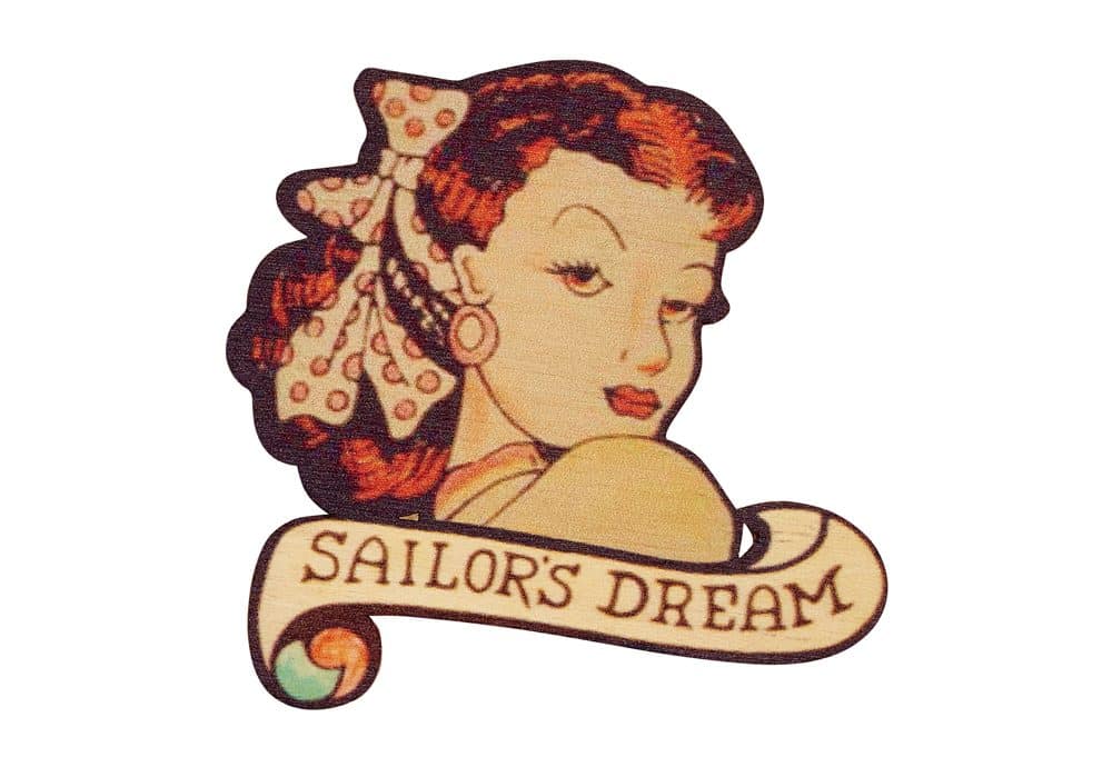 A scan of a broach inspired by famous tattoo artist Jessie Knight, made by Tatty Devine. A woman with red hair looks over her shoulder, with 'sailor's dream' written underneath.