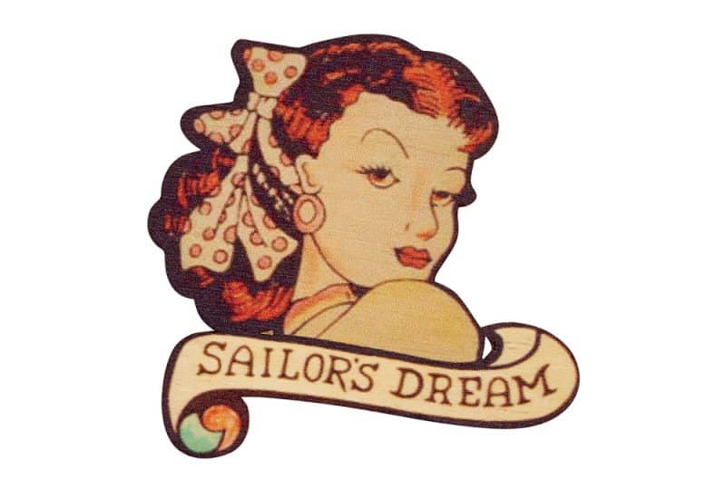 A scan of a broach inspired by famous tattoo artist Jessie Knight, made by Tatty Devine. A woman with red hair looks over her shoulder, with 'sailor's dream' written underneath.