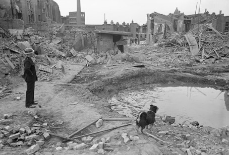 Black and white photo of a man looking at a ruined building with debris all around him. To his right is a dog.