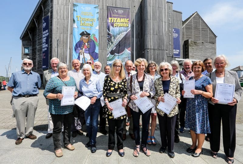 Photo of a group of volunteer standing on Events Square with the Museum in the background. Those in the front row are holding certificates.