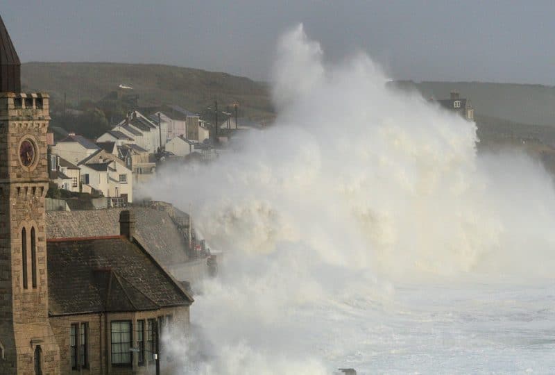 Photo of large waves crashing over the harbour wall in Porthleven, with a church in the foreground.