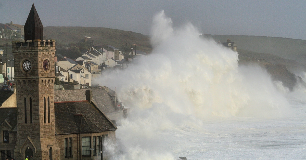 Photo of large waves crashing over the harbour wall in Porthleven, with a church in the foreground.