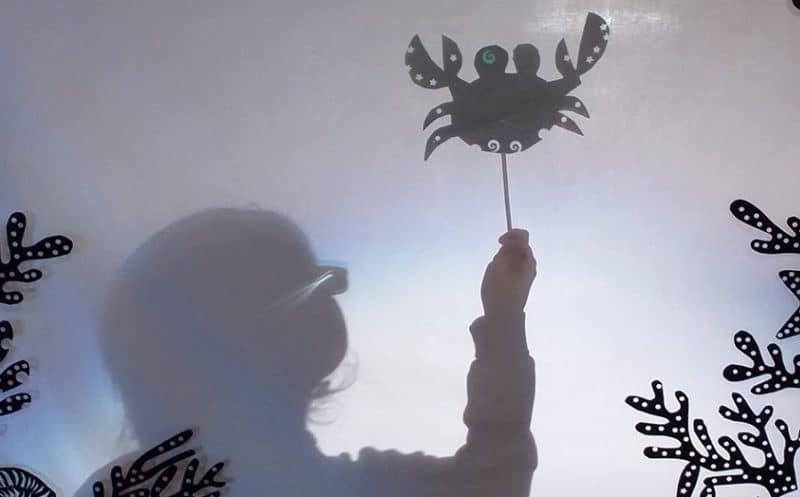 The silhouette of a child holds up a shadow puppet shaped like a crab.