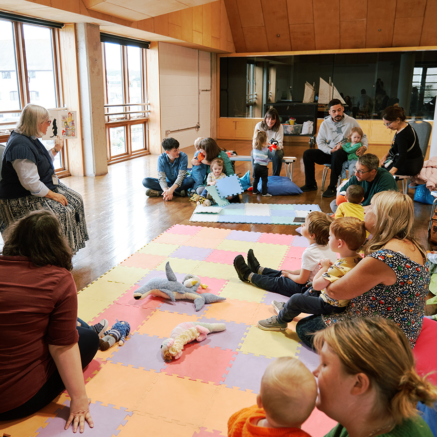 A photo of a lady reading a story to a group of adults and children.