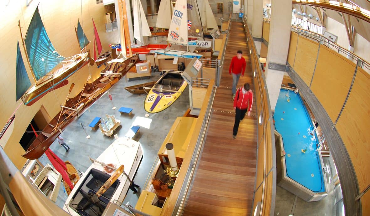 Photo taken from above of two visitors walking down the ramp in the Museum's Main Hall with the flotilla of small boats on the left of the image.