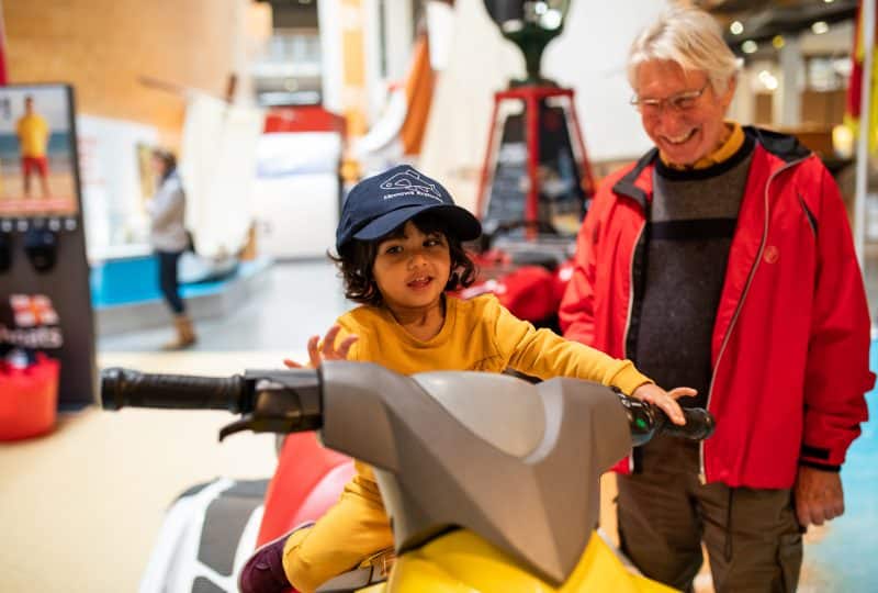 A young boy plays on a static jet ski on display in the Museum while his grandfather smiles at him.
