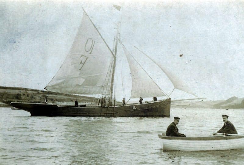 A black and white photo of a Falmouth cutter, with two men rowing past in a small boat in the foreground.