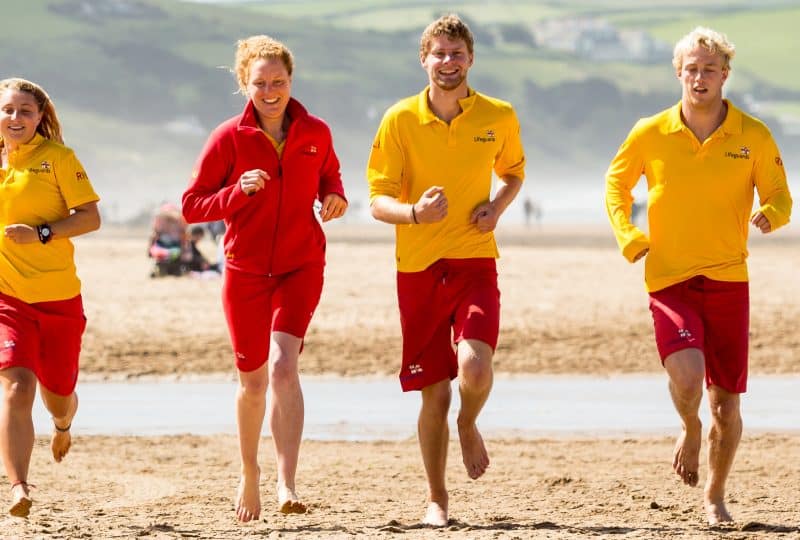 Four lifeguards - two male and two female - run across a sandy beach towards the camera.