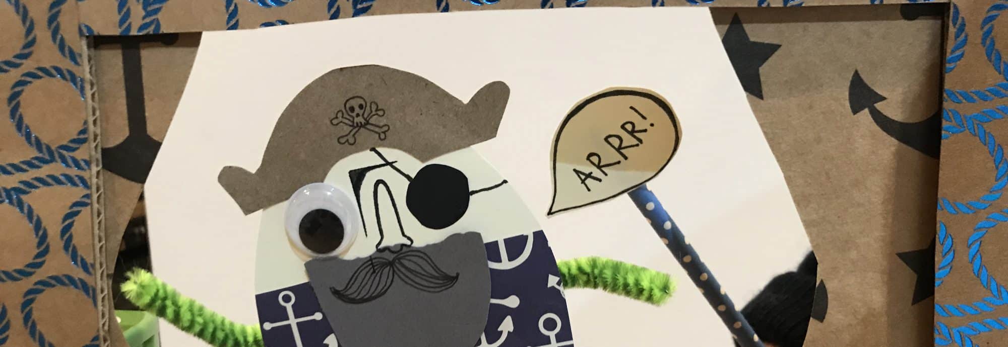 A photo of a pirate finger puppet made from card, pipe cleaners and googly eyes. A speech bubble is held up next to it that says 'Arrr!'