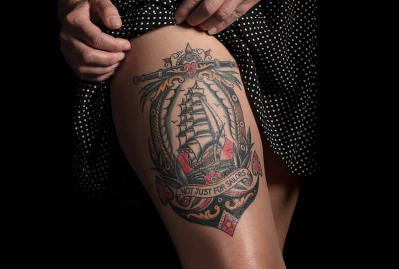 A woman shows a tattoo on her thigh that depicts a sailing ship and has the phrase 'not just for sailors' underneath.