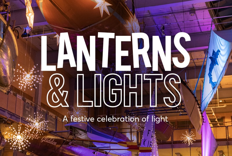 White text in the centre of the image reads 'Lanterns & Lights' and 'A festive celebration of light'. In the background, warm lights hang from the ceiling of the Museum's Boat Hall as well as fabric banners.