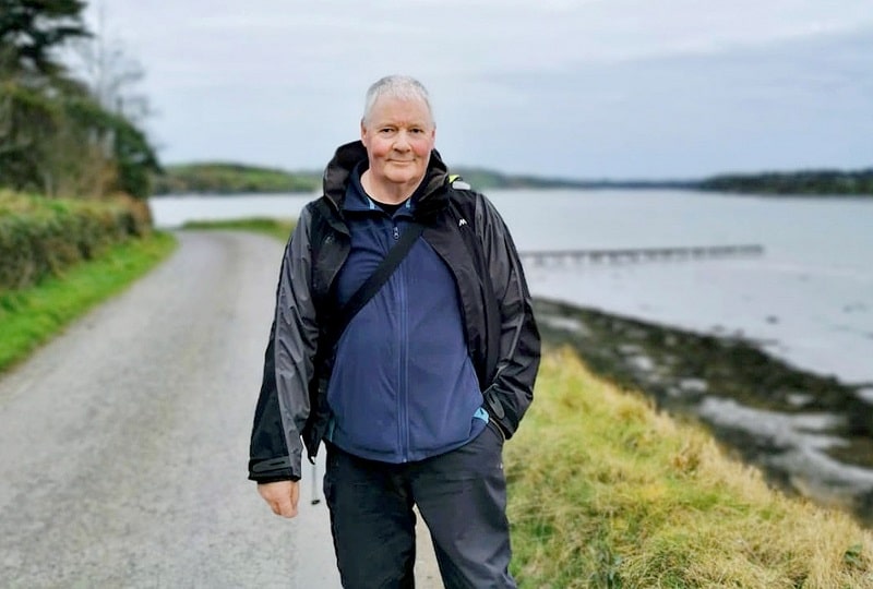 Kevin O'Farrell smiles for the camera. He is walking along a road with a river to his left.