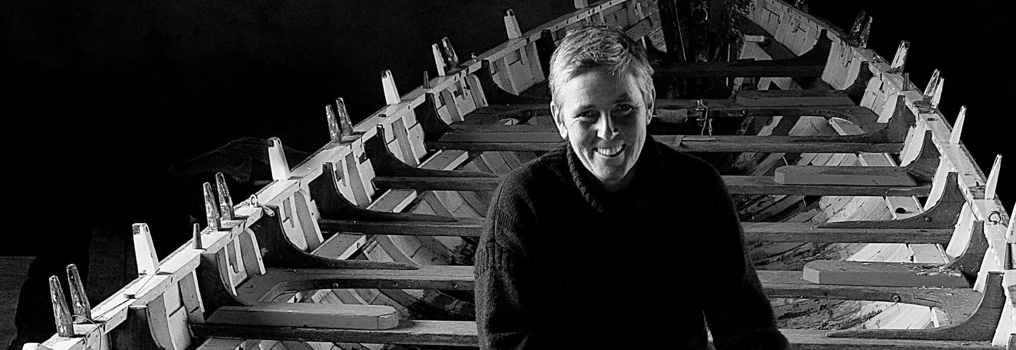 A black and white image of Gail McGarva smiling for the camera in front of a wooden boat.