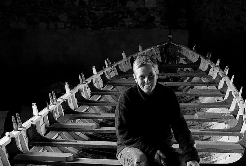 A black and white image of Gail McGarva smiling for the camera in front of a wooden boat.