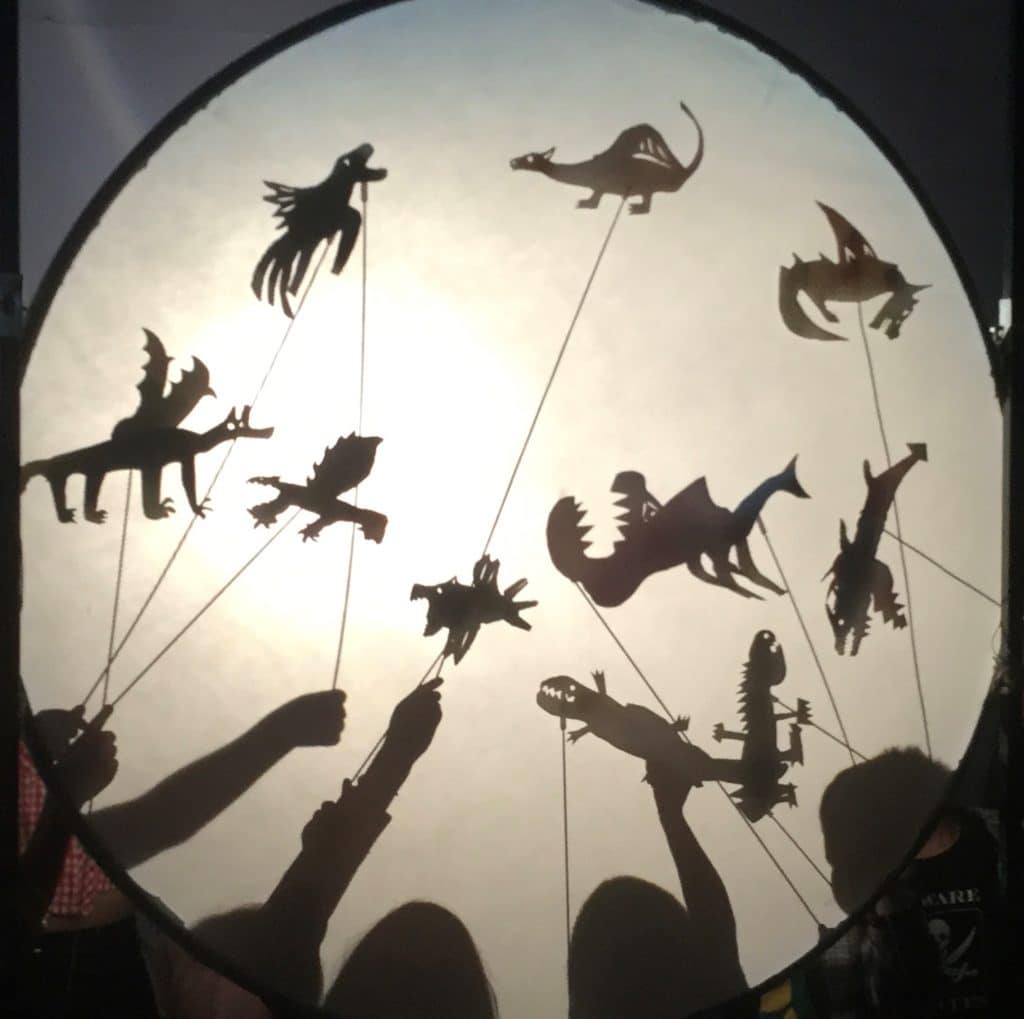 Cut-out dragons and dinosaurs are held up so that they are silhouetted against a white background.