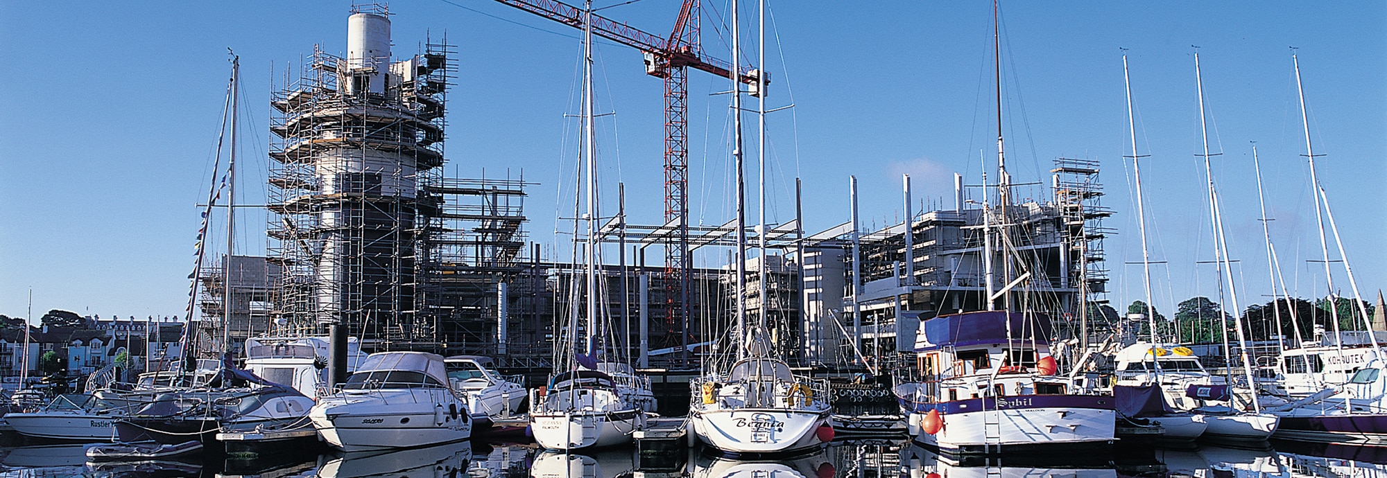 In the foreground, a line of yachts are moored up in Falmouth harbour. In the background, the Museum is covered in scaffolding, in the middle of being built.