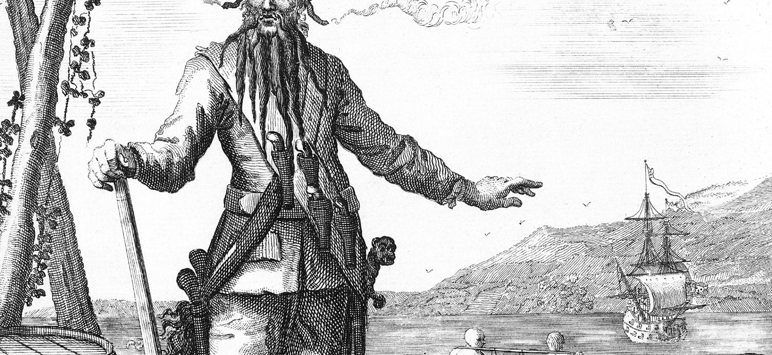 Black and white drawing of the pirate Blackbeard. In the background, two men carry a barrel between them and a ship is anchored out to sea.