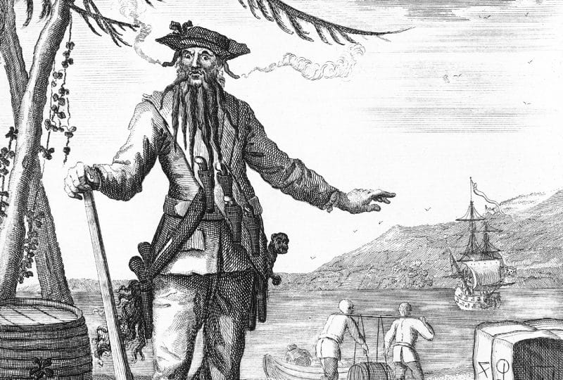 Black and white drawing of the pirate Blackbeard. In the background, two men carry a barrel between them and a ship is anchored out to sea.