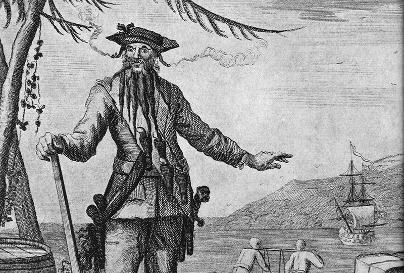 Black and white illustration of the pirate Blackbeard. In the background, two men carry a barrel between them and a ship is anchored in the harbour.
