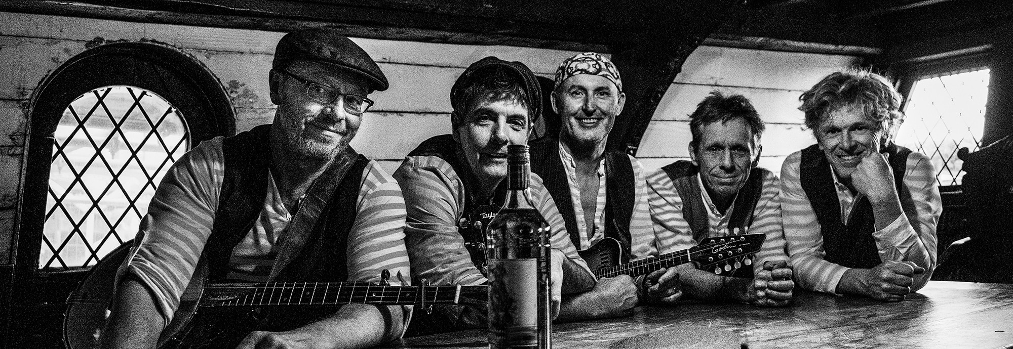 Black and white image of five men sat at a wooden table, smiling at the camera. Two wear caps, one wears a bandana. Two are holding guitars. A large bottle sits on the table in front of them.