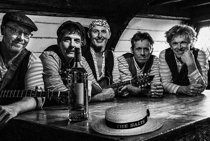 Black and white image of five men sat at a wooden table, smiling at the camera. Two wear caps, one wears a bandana. Two are holding guitars. A large bottle and a straw cap with a band saying 'The Salts' sit on the table in front of them.