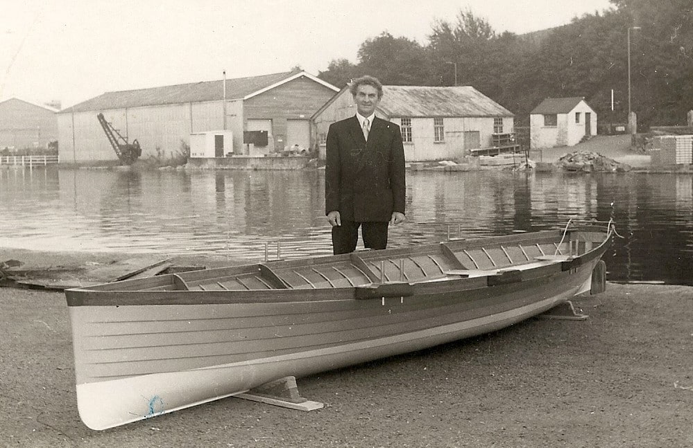 A black and white photo of a man, called Victor Angove, stood next to a wooden boat called Angof. The photo was taken at the launch of the boat.