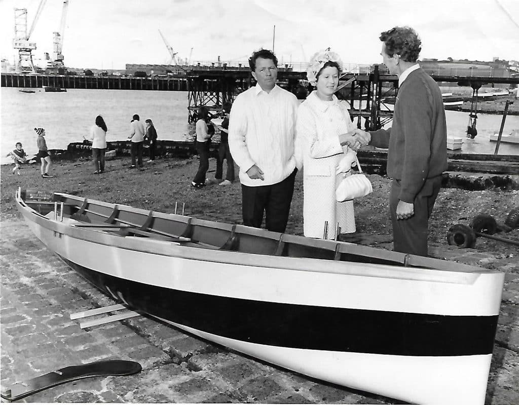 A black and white photo of three people next to a wooden boat. The photo was taken on Falmouth foreshore with Falmouth Docks in the background. The people in the photo are Doug Thomas, Youth Club Chairman, Clive Sampson is seen with Mrs Jean Thomas, Former Mayoress of Penryn and Victor Angove, who designed and built the boat.