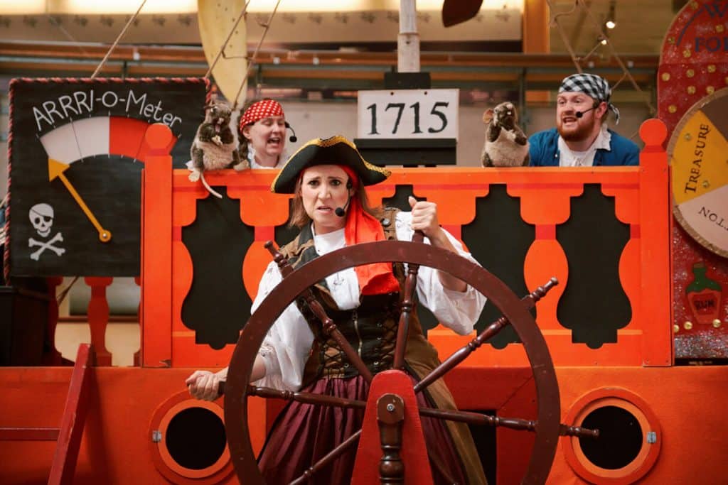 A picture taken of the interactive theatre show, A Pirate's Life For Me!, at the National Maritime Museum Cornwall. Three pirates are on a stage constructed in the style of a pirate ship. A pirate in the foreground is at the helm while two pirates are behind her operating rat hand puppets.