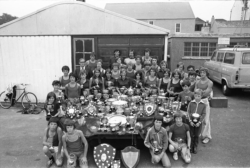 A black and white photo of the members of Penryn Rowing Club with a large array of the trophies they have won.