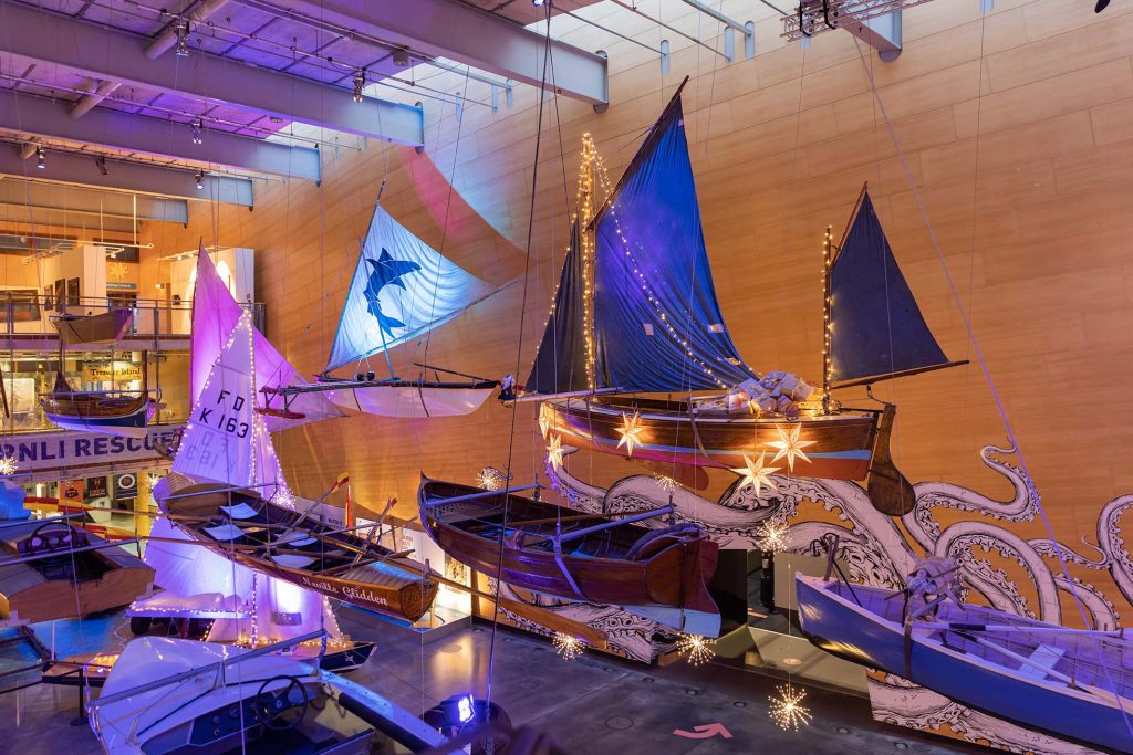 Photo of the Main Hall at the Museum, with warm lights draped across several of the boats suspended from the ceiling.
