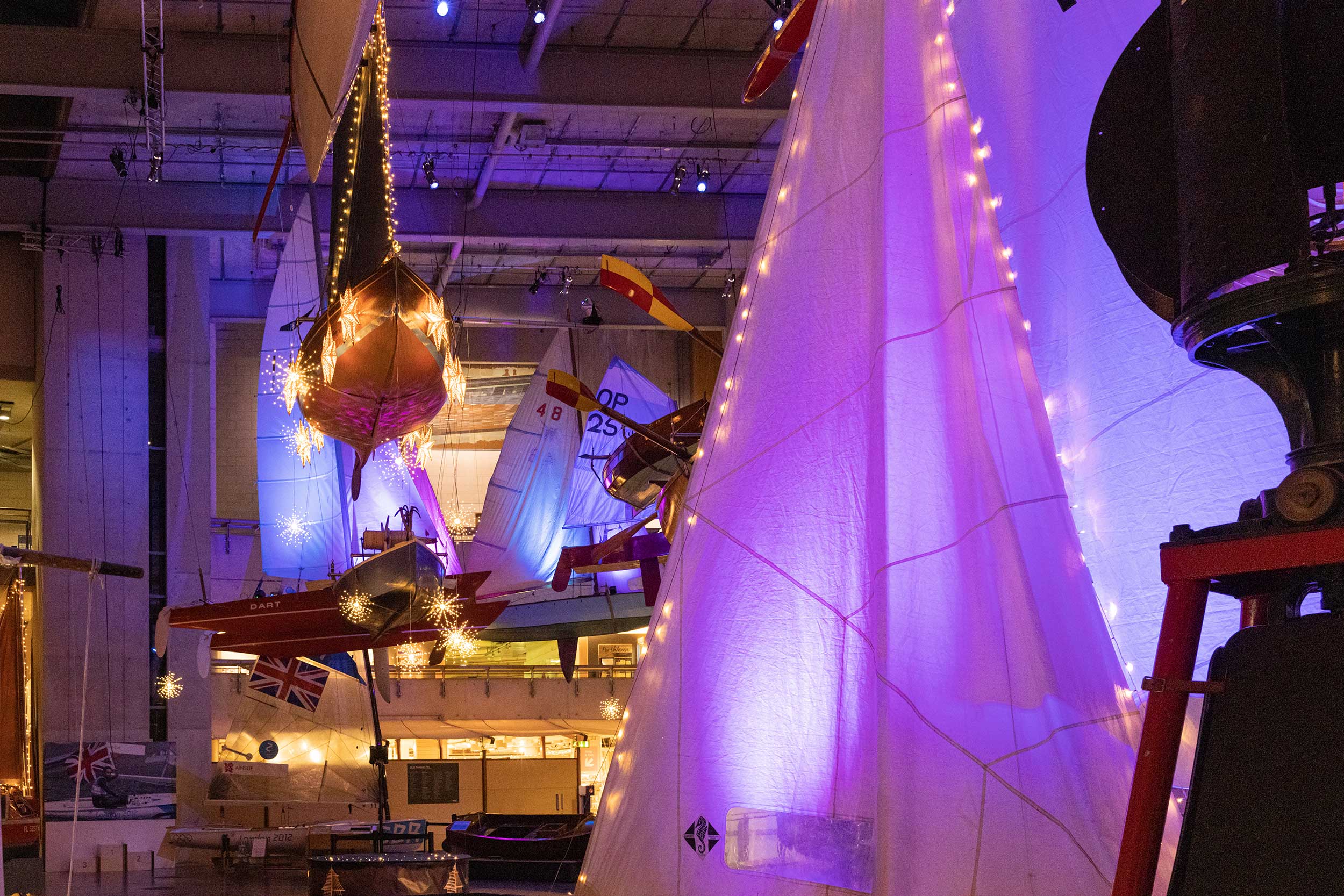 Photo looking up at the boats suspended from the ceiling of the Museum's Boat Hall, draped in warm lights.