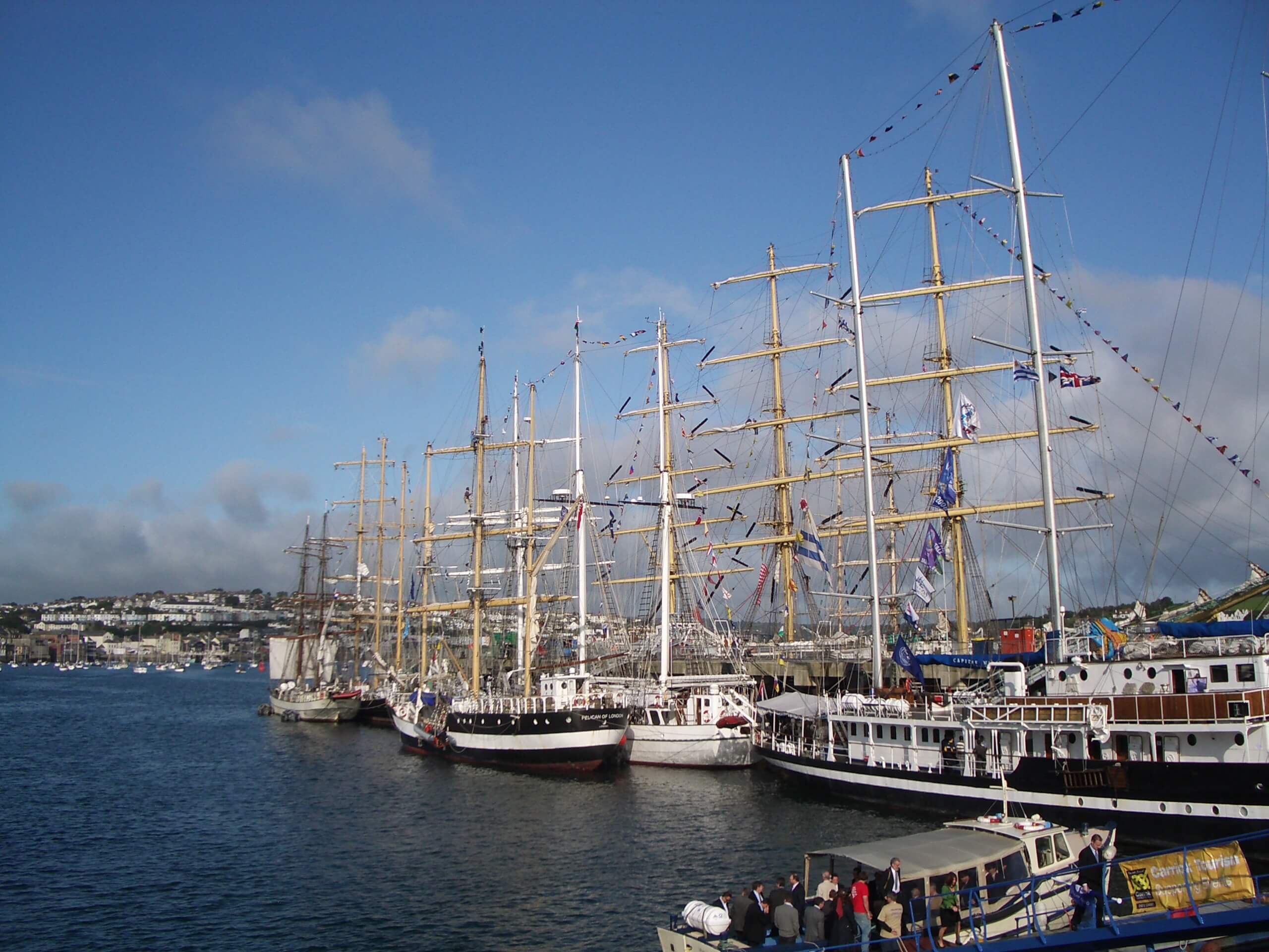 A photo of several tall ships stationed alongside County Wharf, at A&P Falmouth, in 2008. The photo is taken on a cloudy day with blue skies and calm harbour waters.