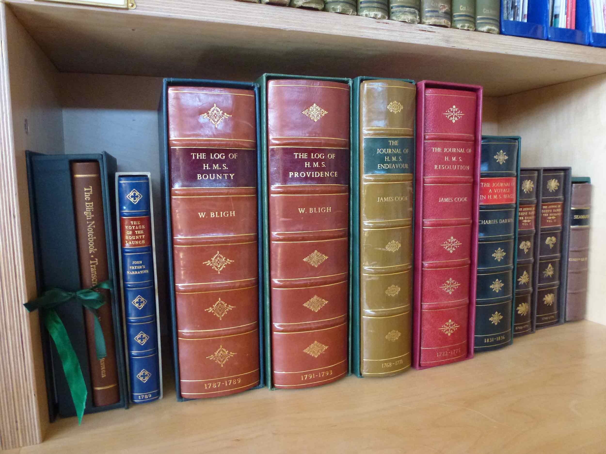 A series of books on display in the Bartlett Library, including the 'Log of H.M.S. Bounty' and 'the Journal of H.M.S. Endeavour'.