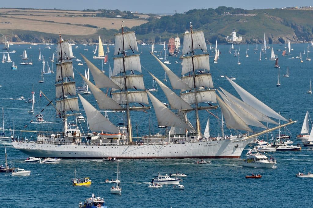 A photo of the Polish Tall Ship Dar Mlodziezy at the Falmouth Tall Ships Parade of Sail. The large white sailing ship is pictured on a sunny day surrounded by dozens of smaller vessels as she sails in Falmouth Bay.