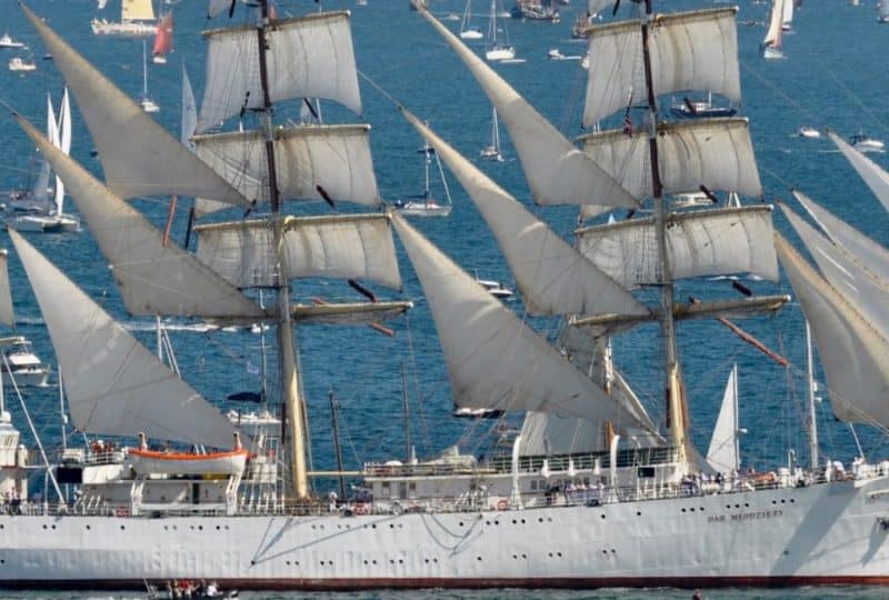 A photo of the Polish Tall Ship Dar Mlodziezy at the Falmouth Tall Ships Parade of Sail. The large white sailing ship is pictured on a sunny day surrounded by dozens of smaller vessels as she sails in Falmouth Bay.