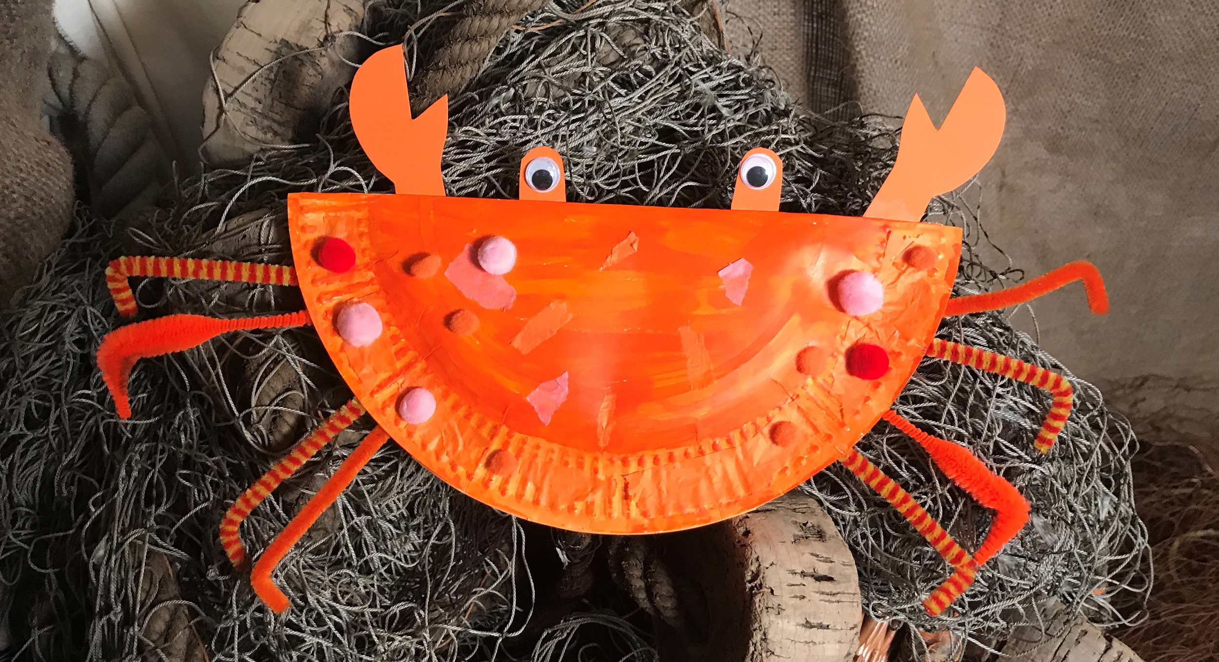 A photo of a crab made from a paper plate, pom poms, googly eyes and pipe cleaners.