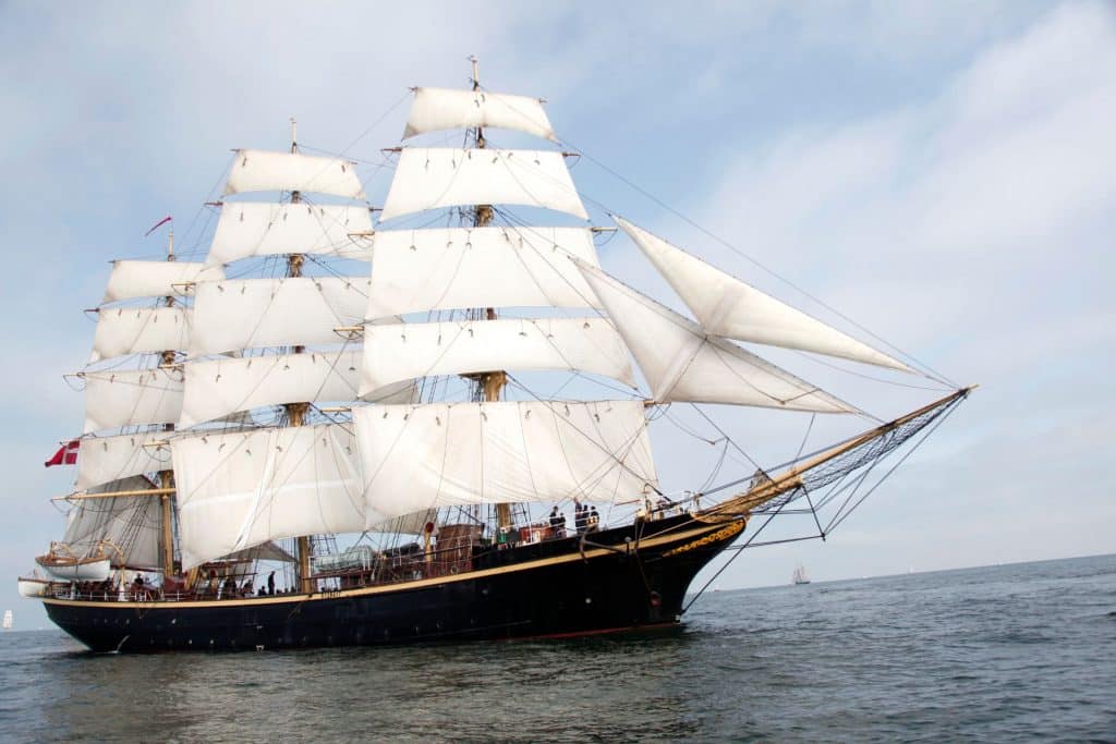 A photo of the tall ship Georg Stage. The large three-masted sailing ship has a black steel hull and white sails. Pictured on a calm sunny day.